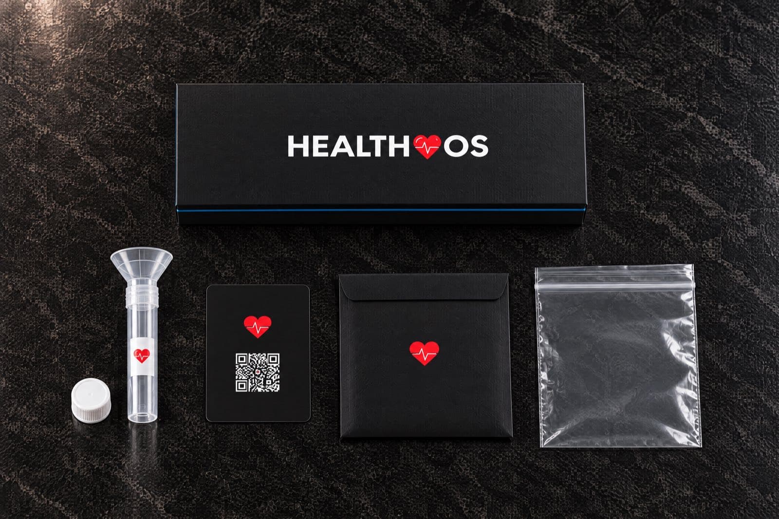 Seen from above on dark marble: the closed black HealthOS box, and below it the saliva tube with its funnel and cap, the black kit card with a QR code, a black envelope with the red heart and a clear specimen bag