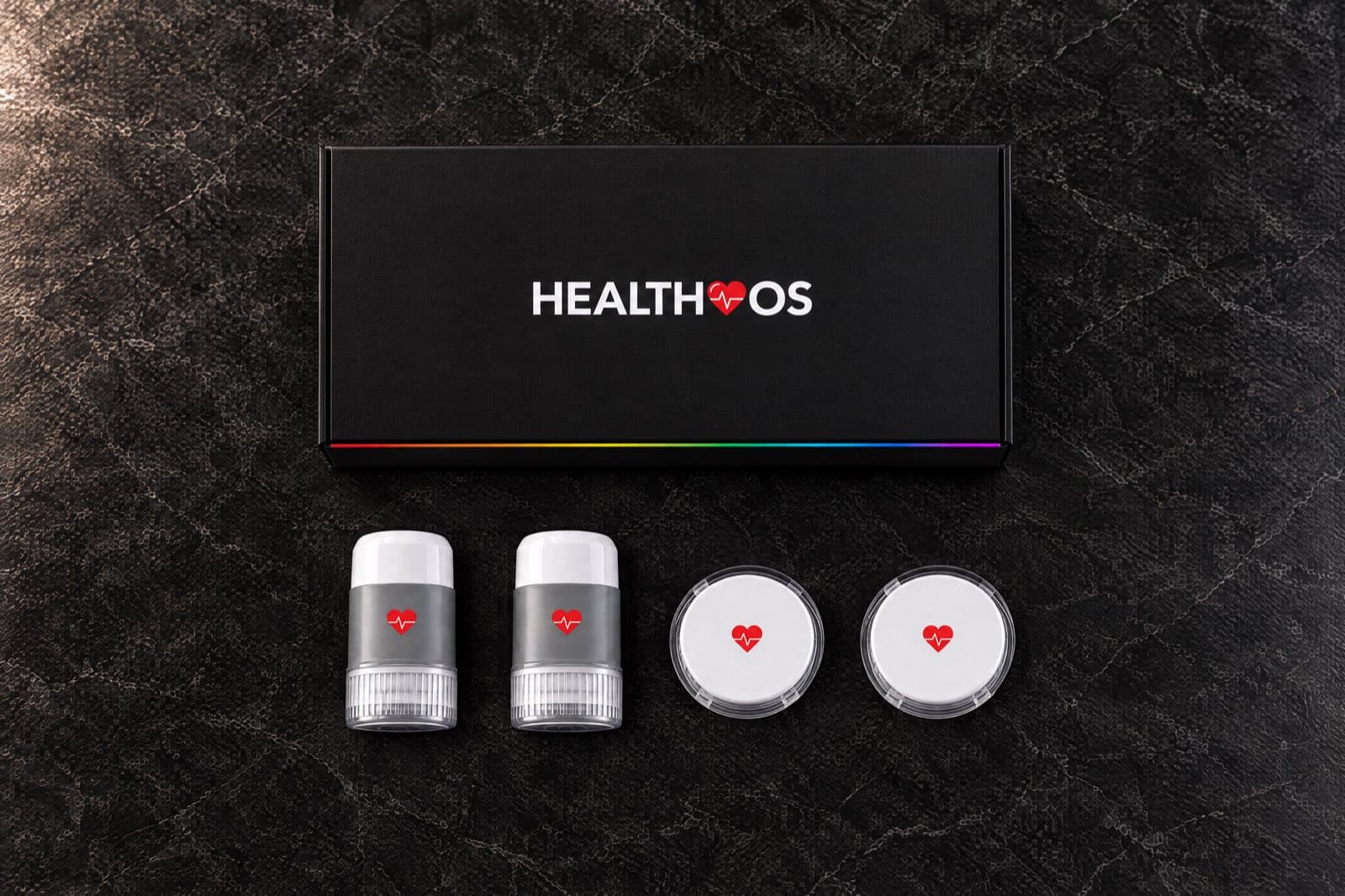 Seen from above on dark marble: the closed black HealthOS Sensor box with a rainbow edge, and below it two grey-and-white applicators and two white sensors with the red HealthOS heart